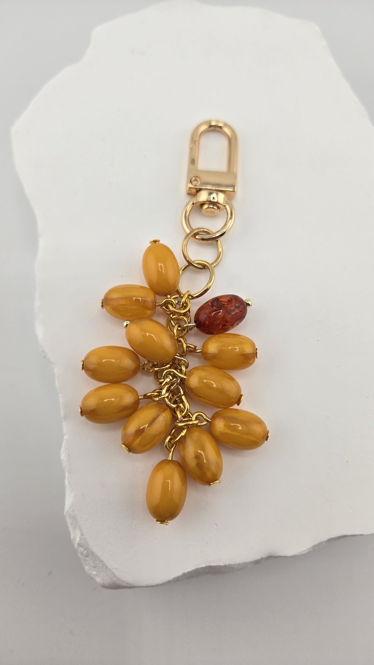 Butterscotch Bag Charm, a cluster of butterscotch-colored oval beads and one dark amber-colored bead on gold-plated chain with a gold-tone swivel clip, on a white stone