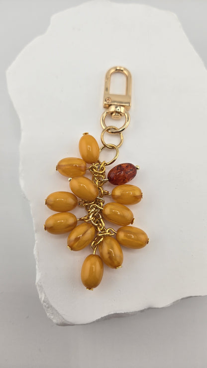Butterscotch Bag Charm, a cluster of butterscotch-colored oval beads and one dark amber-colored bead on gold-plated chain with a gold-tone swivel clip, on a white stone