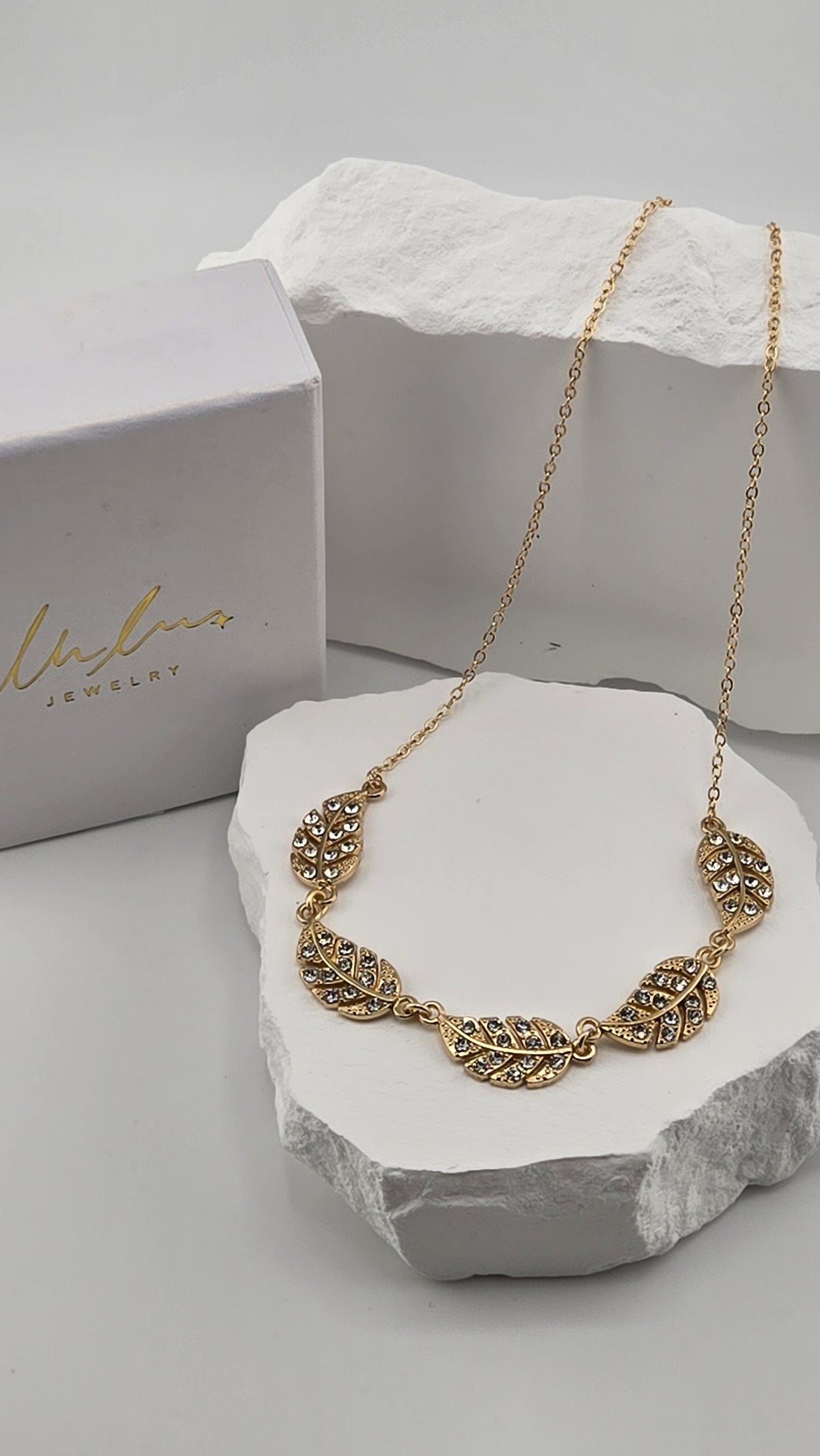 Feather Garland Necklace laid in a curve on a white stone next to a LuluTouch gift box