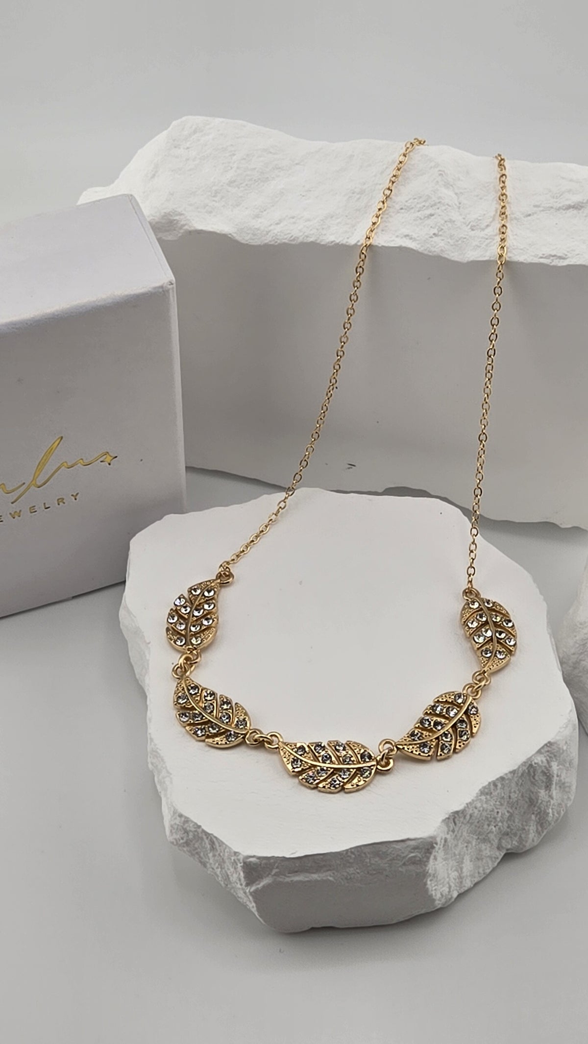 Feather Garland Necklace, five linked gold-plated feathers set with small clear stones on a cable chain, resting on white stones beside a LuluTouch box