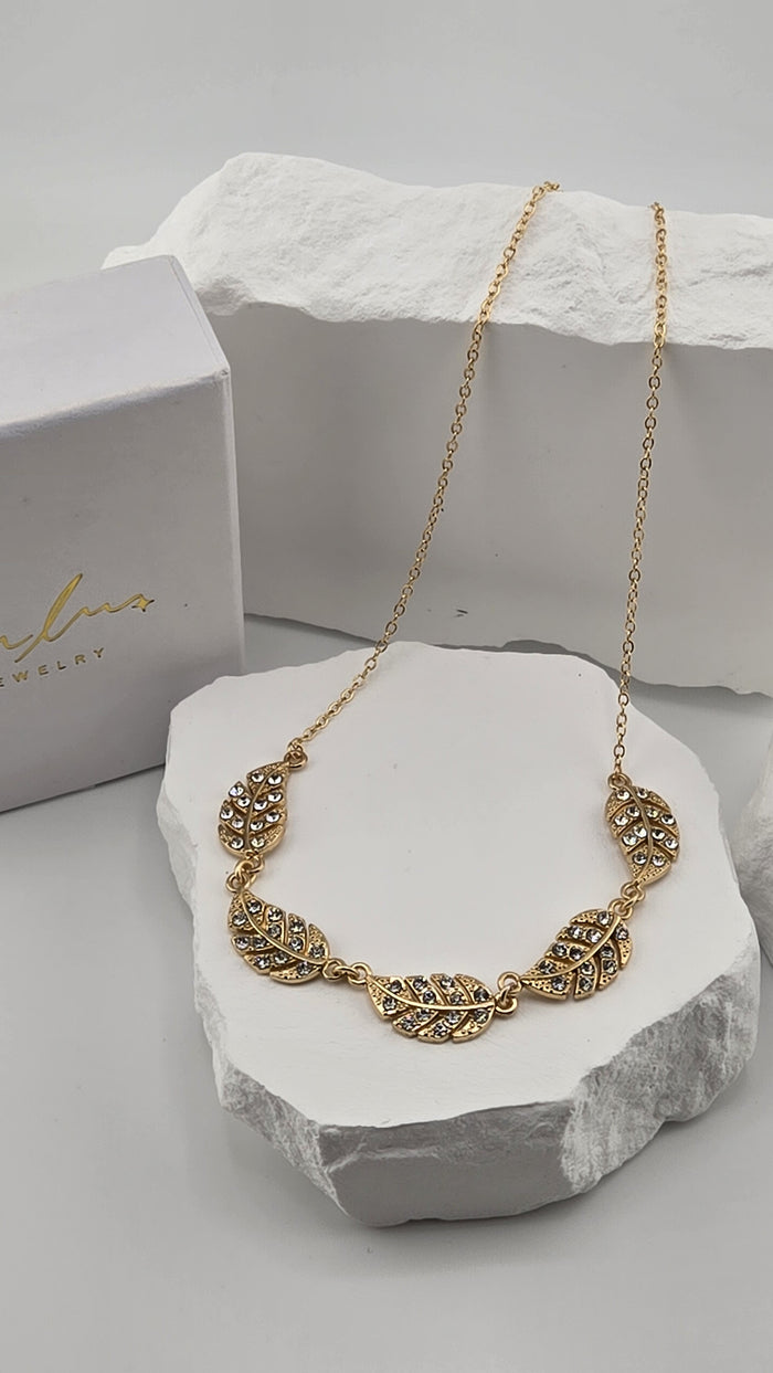 Feather Garland Necklace, five linked gold-plated feathers set with small clear stones on a cable chain, resting on white stones beside a LuluTouch box