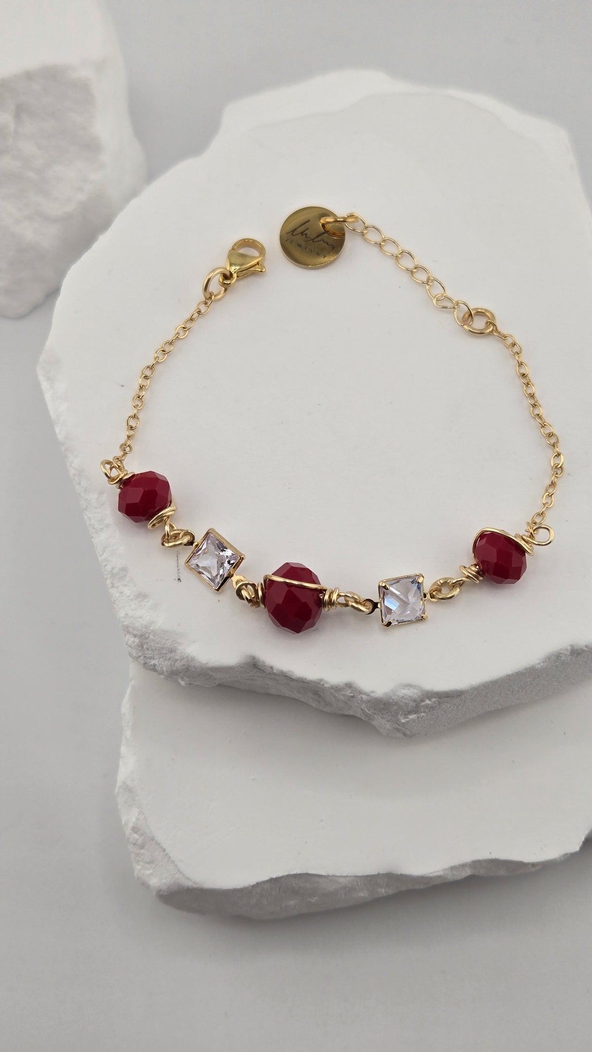 Poppy Facet Bracelet, three deep red faceted beads wrapped in gold-plated wire with two square clear stones on a gold-plated chain, on a white stone