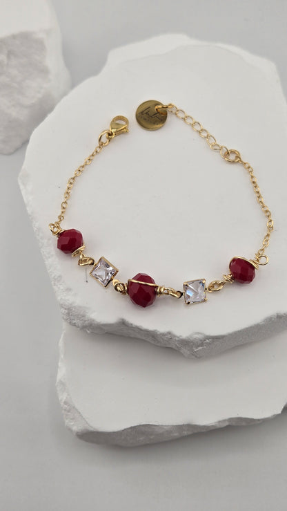 Poppy Facet Bracelet, three deep red faceted beads wrapped in gold-plated wire with two square clear stones on a gold-plated chain, on a white stone