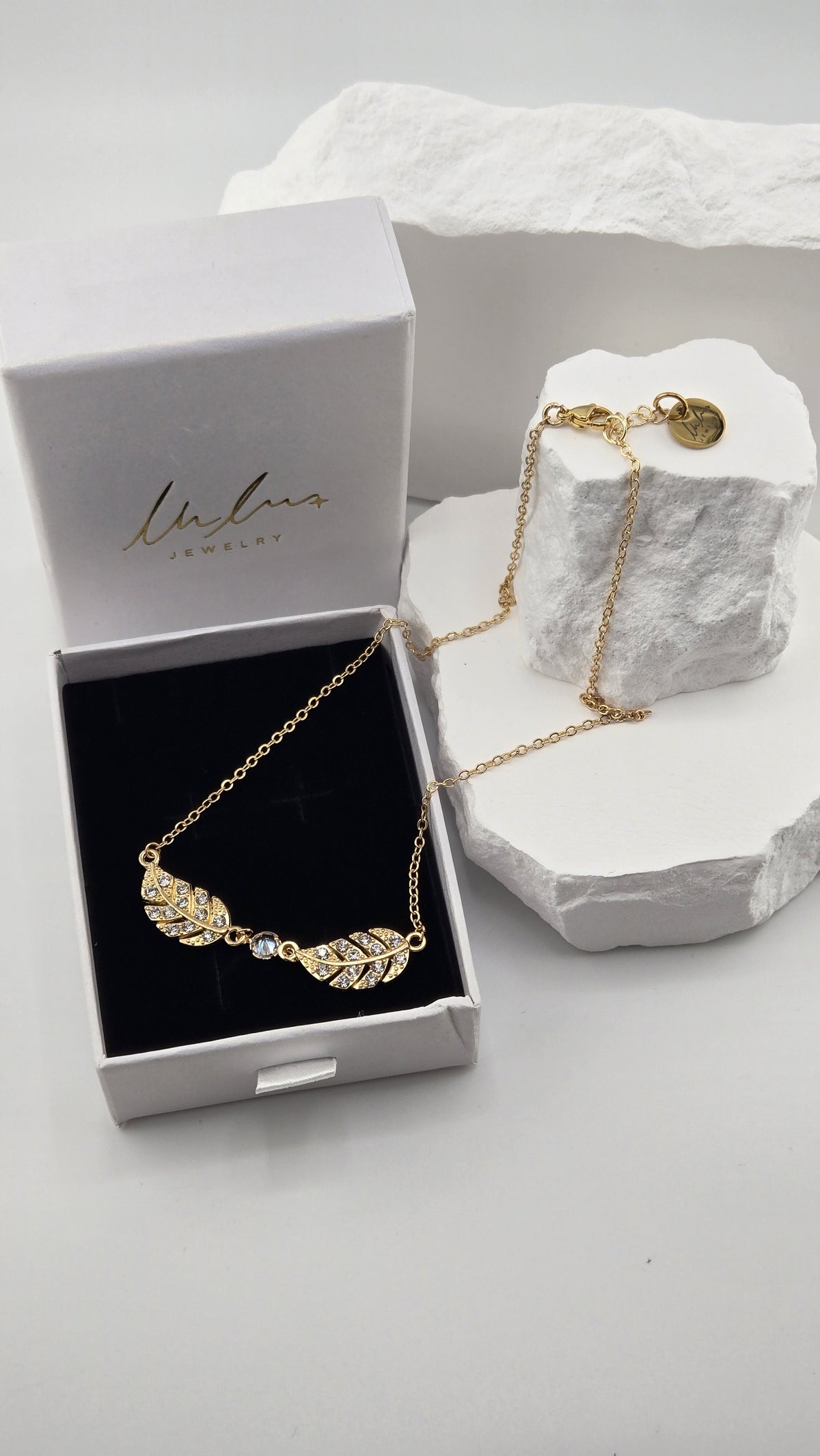 Twin Plume Necklace, two 18K gold-plated plume pendants set with clear stones around a round clear center stone, in a LuluTouch gift box on white stones
