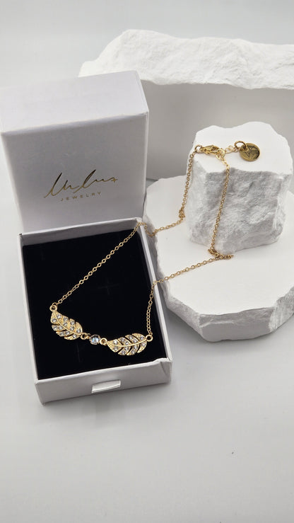 Twin Plume Necklace, two 18K gold-plated plume pendants set with clear stones around a round clear center stone, in a LuluTouch gift box on white stones