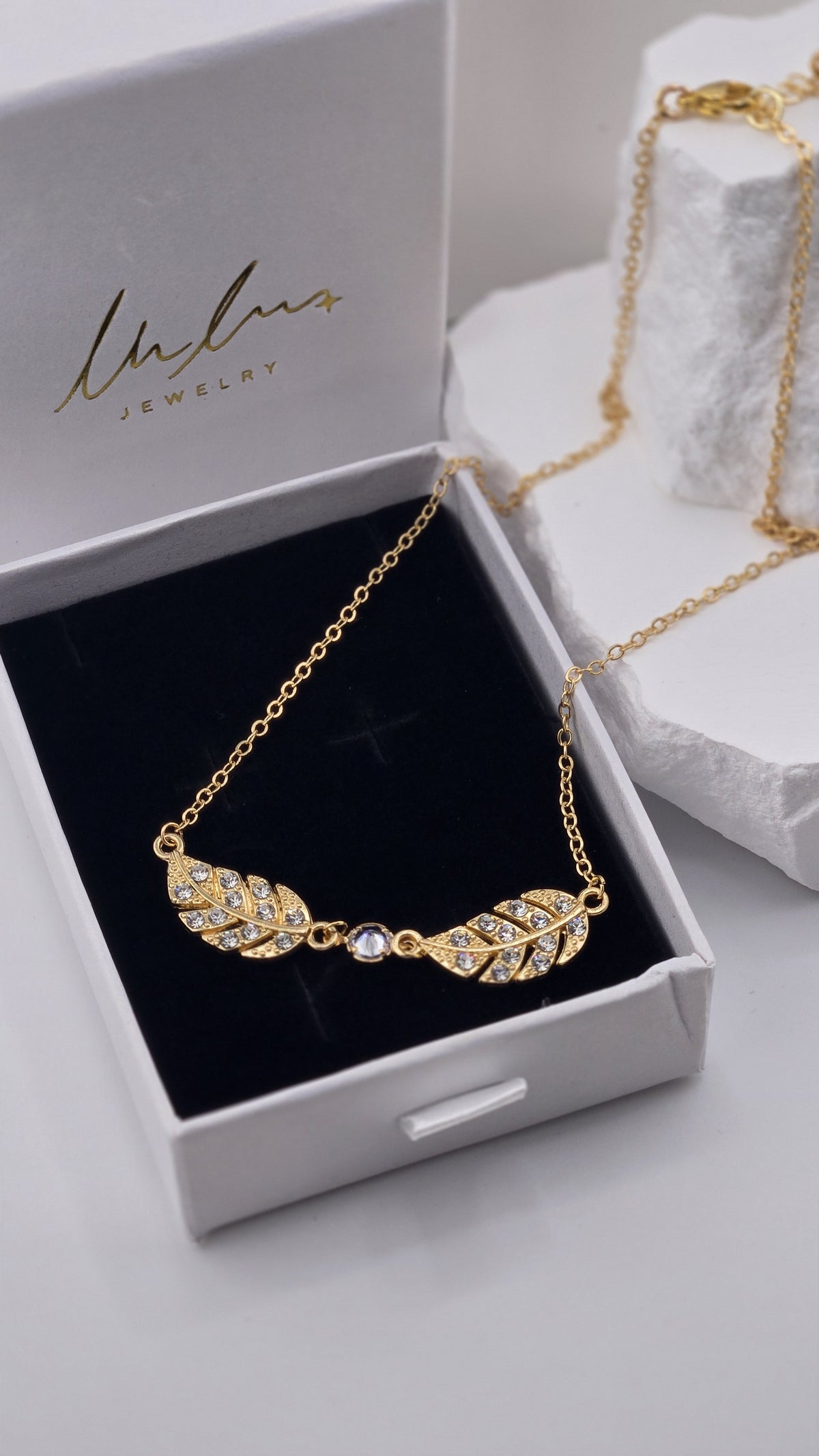 Close-up of the Twin Plume Necklace pendants, two gold-plated plumes with small clear stones and a clear center stone on black velvet