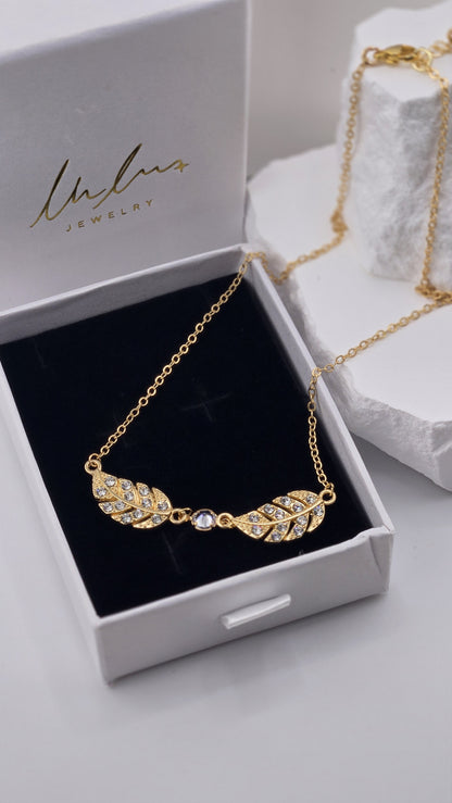 Close-up of the Twin Plume Necklace pendants, two gold-plated plumes with small clear stones and a clear center stone on black velvet