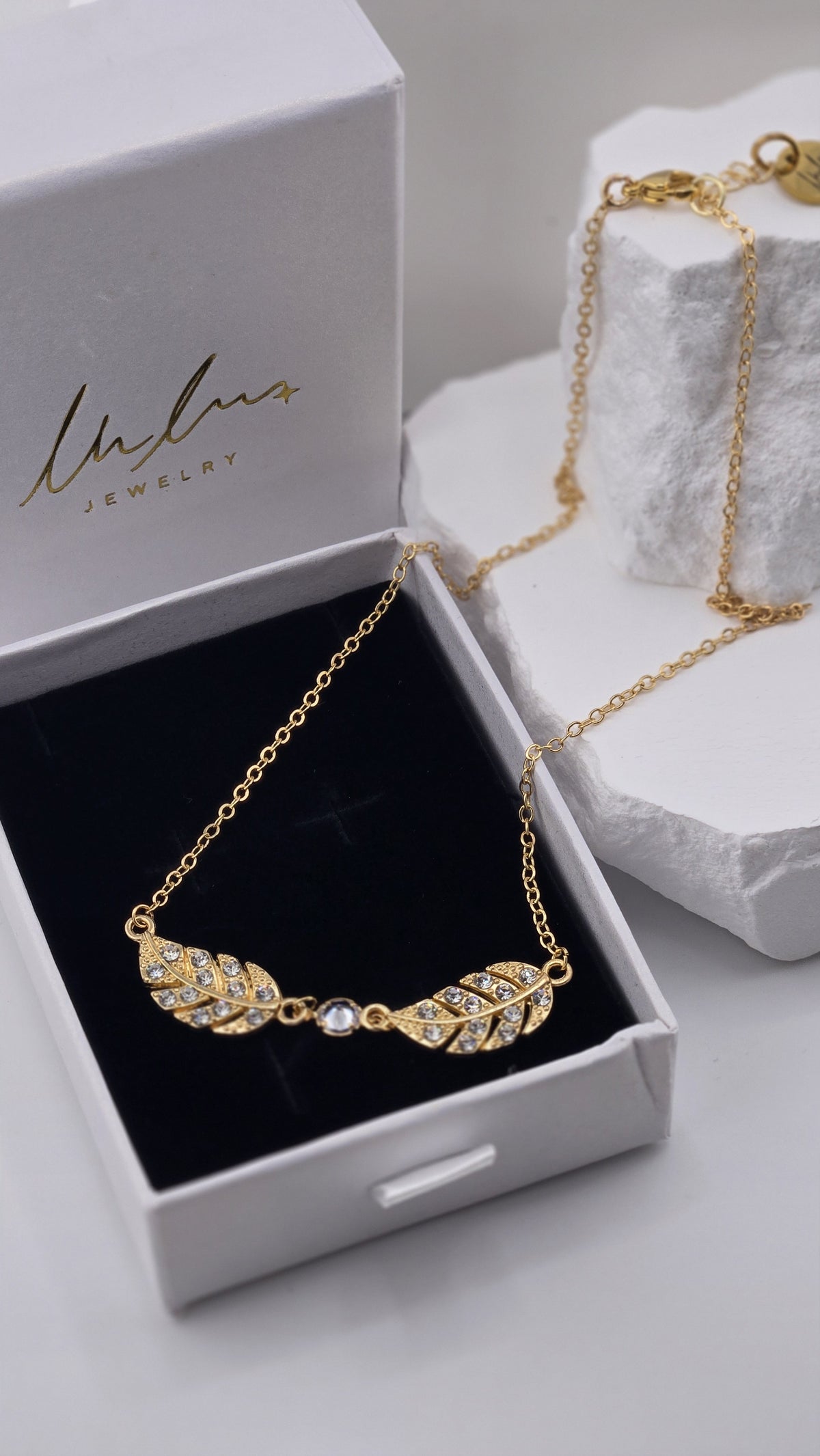 Twin Plume Necklace pendant detail with clear stones set in two gold-plated plumes, chain draped over a white stone