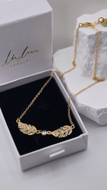 Twin Plume Necklace pendant detail with clear stones set in two gold-plated plumes, chain draped over a white stone