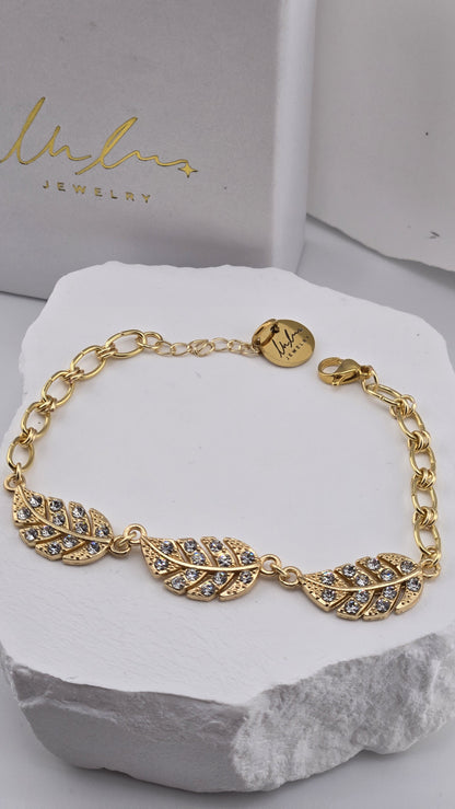 Wingtip Bracelet laid open on a white stone, three feathers with clear stones and a gold-plated link chain