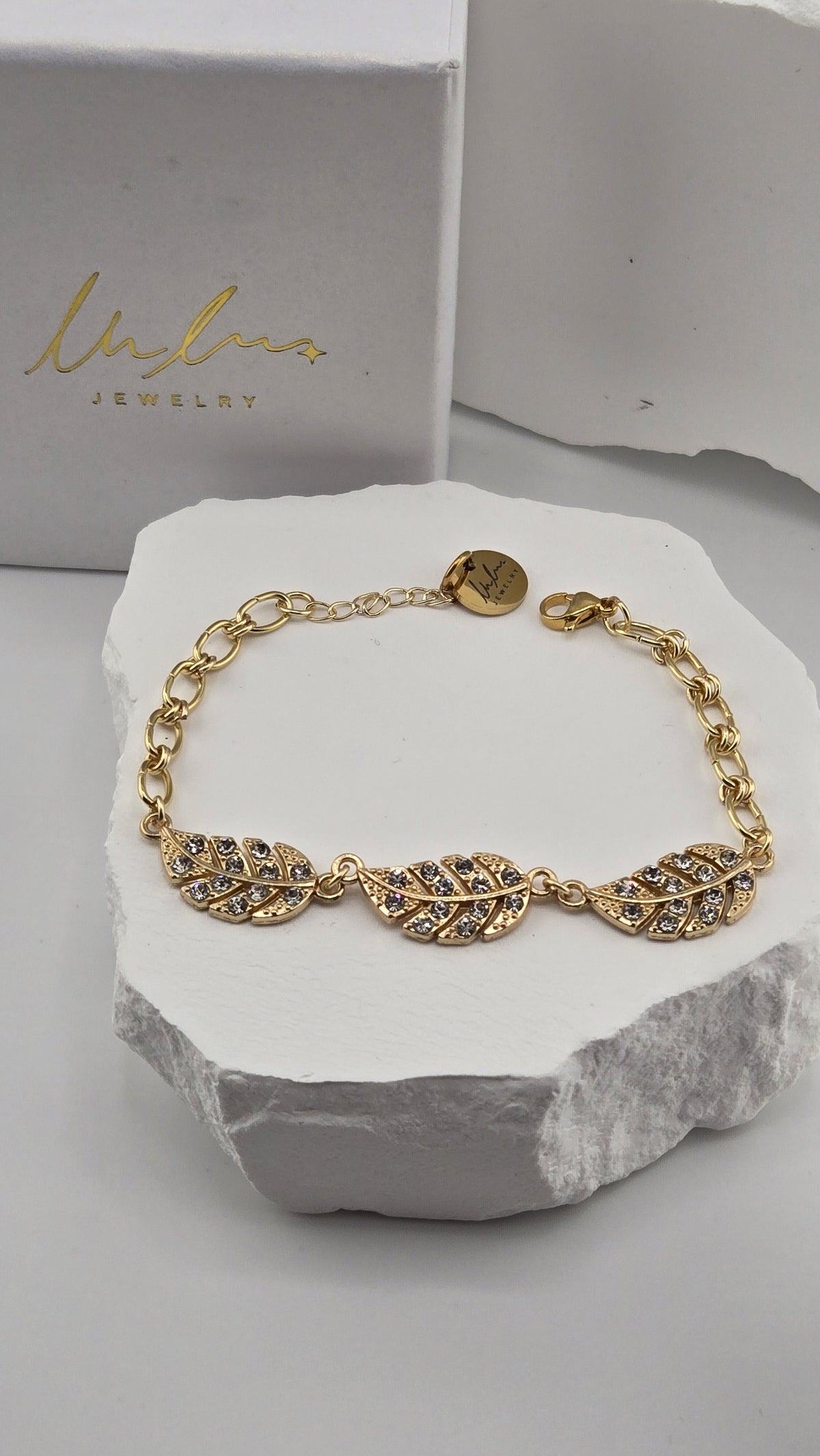 Wingtip Bracelet, three gold-plated feathers set with small clear stones on a chunky link chain, resting on a white stone beside a LuluTouch box